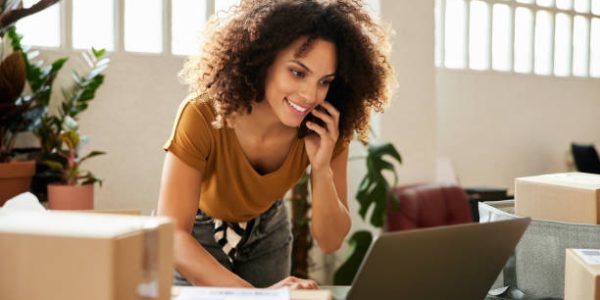 Young female talking on smart phone while using laptop. Confident businesswoman is working at home office. She is having curly hair.